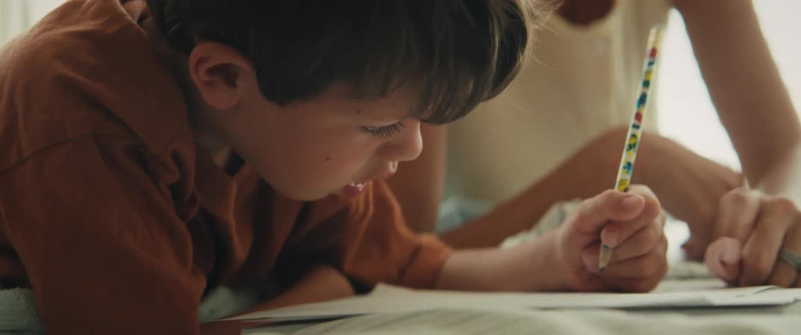 A loving mother gently encourages her young son as he lies down and carefully draws with a pencil at home