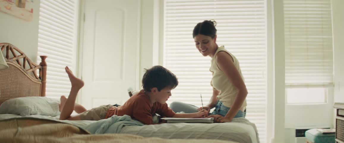 A smiling mother helps her young son with his drawing as they spend quality time together on a bed in a bright bedroom