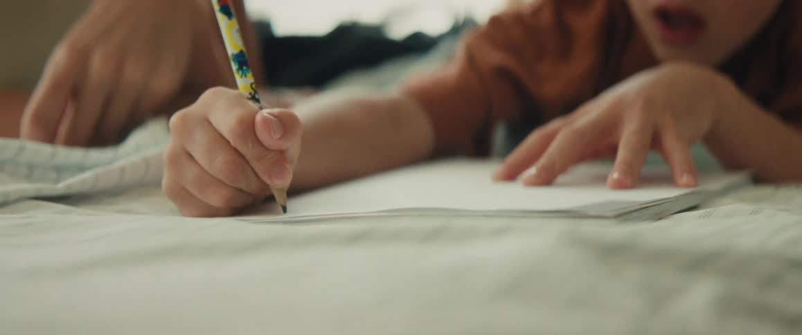 A close-up of a young boy intensely focused on drawing with a pencil while his mother's gentle hand rests on his shoulder