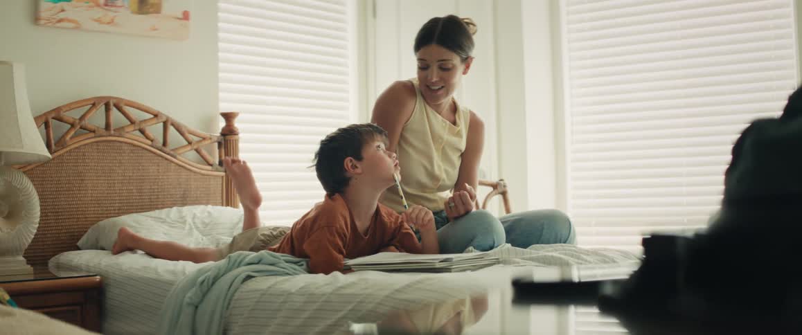 A young boy lies on his stomach on the bed, drawing in a notebook, while his mother sits beside him offering loving encouragement