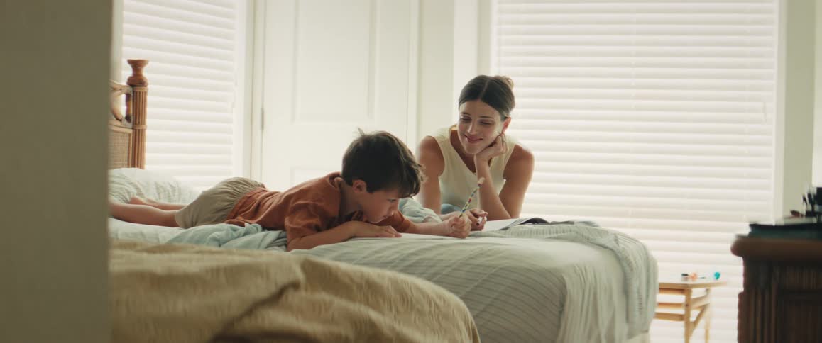 A loving mother helps her young son with his homework as they relax together on the bed in a sunlit bedroom