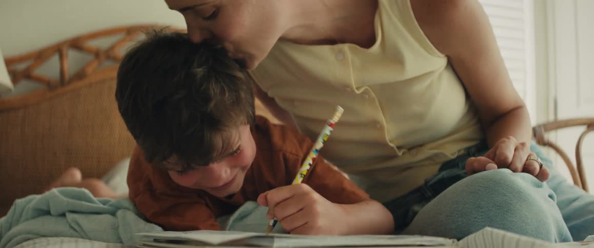 Close-up of a smiling boy drawing in a notebook on his bed as his mother affectionately kisses his head