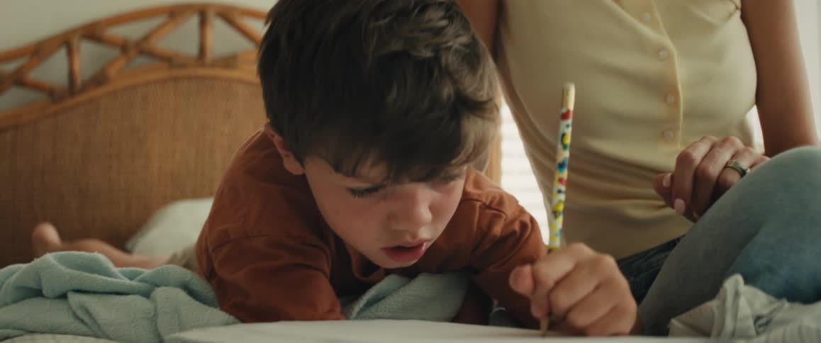A young boy carefully writes in a notebook while his mother sits beside him on a bed and helps him learn