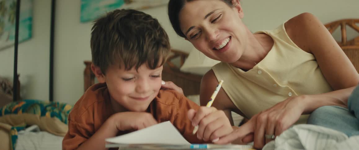 A smiling mother helps her young son draw in a coloring book while they relax on a bed in a brightly lit room
