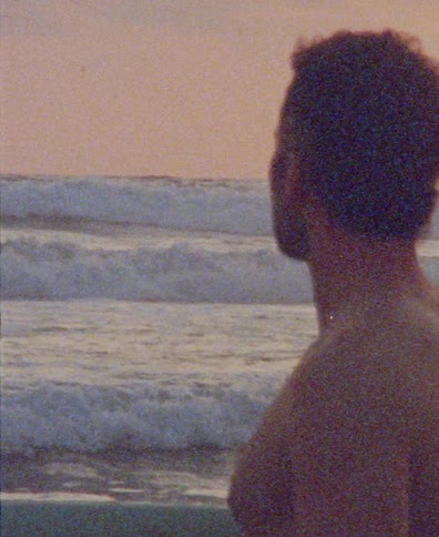 A grainy, vintage-style shot of a man from behind watching the ocean waves at sunset