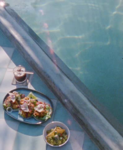 A relaxing poolside scene features a plate of tacos, a side of guacamole, and a drink under the bright sun