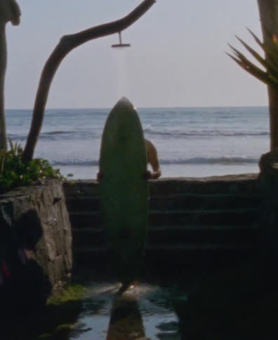 A person rinses off their surfboard under a beachside shower with an ocean view