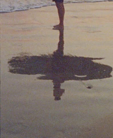 Grainy, vintage-style footage of a person's reflection on the wet sand at the ocean's edge during sunset