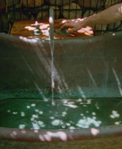 A hand turns on a vintage tap, filling a rustic copper bathtub with water in a sun-dappled outdoor setting