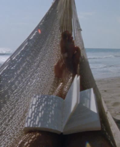From a first-person perspective, an open book's pages flutter in the wind as a person relaxes in a hammock on a sandy beach by the ocean