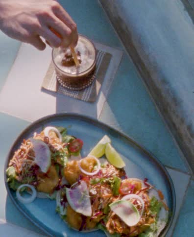 A person stirs a refreshing iced drink next to a delicious plate of gourmet tacos on a sunny day