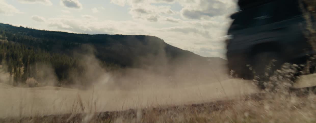 An off-road SUV speeds down a dusty country road, kicking up a large cloud of dust in its wake