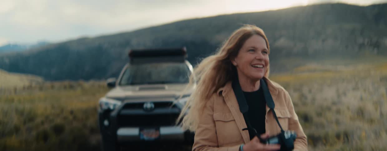 A smiling female photographer walks through a grassy landscape in slow motion before raising her camera to capture the view