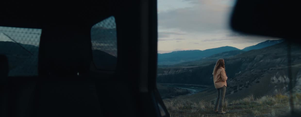 A woman stands on a grassy hillside at twilight, viewed from inside her vehicle, as she looks out over a vast mountain valley and a winding river