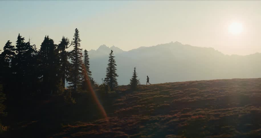 Aerial view of a silhouetted trail runner jogging across a mountain ridge during a hazy sunrise, with a vast mountain range in the background