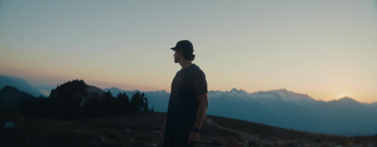A silhouetted hiker walks to the edge of a mountain ridge and pauses to admire the majestic mountain range at sunrise in slow motion