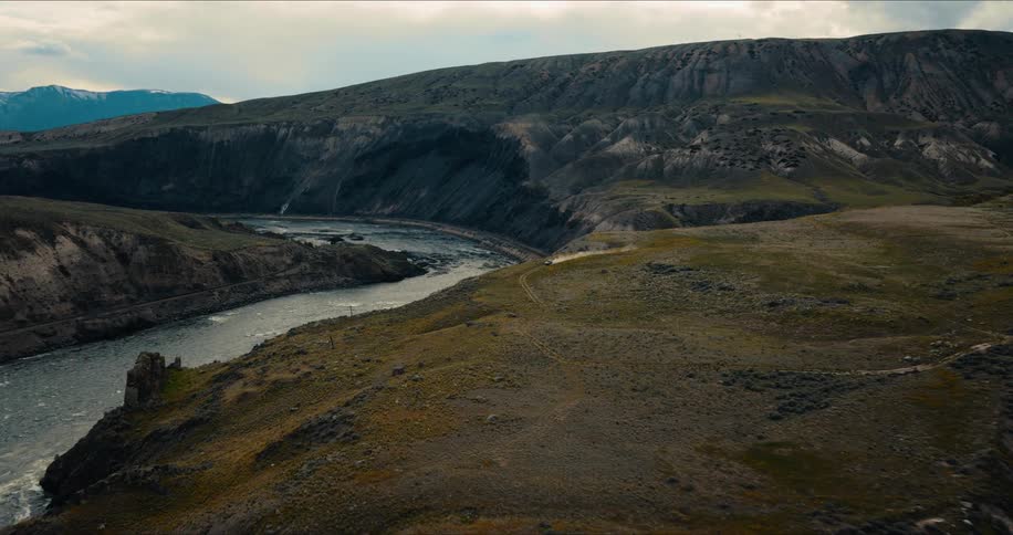 An aerial view shows a white SUV driving along a winding dirt road on a clifftop next to a powerful river in a vast canyon