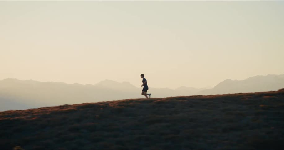 A silhouetted trail runner jogs along a scenic mountain ridge at sunrise, captured in a cinematic slow-motion tracking shot