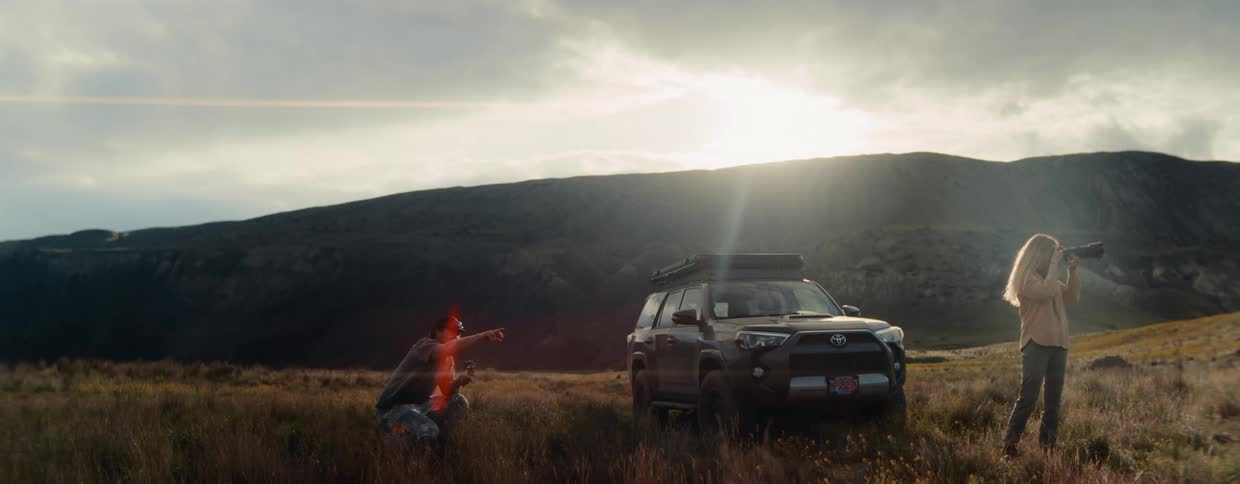 An adventurous couple stops their SUV in a grassy mountain valley to fly a drone and take photographs as the sun sets