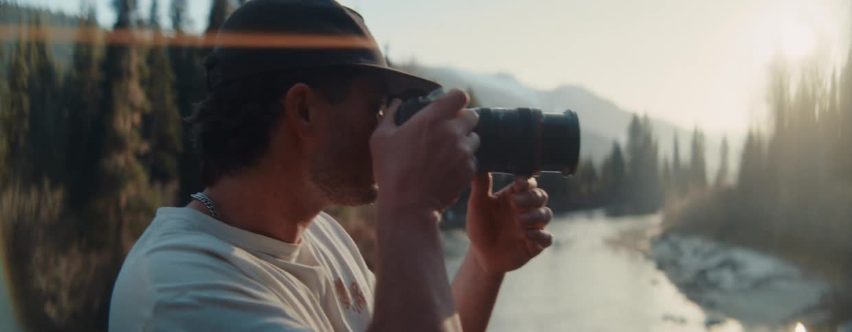 A young male photographer in a hat takes pictures of a scenic river and mountains during golden hour