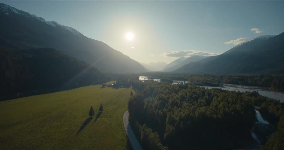 A majestic aerial ascent reveals a beautiful sunlit mountain valley with a winding river, dense forest, and lush green meadow