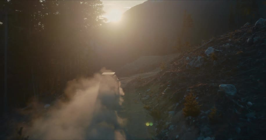 An off-road SUV with a rooftop tent kicks up a cloud of dust as it drives along a remote mountain road into the golden sunrise