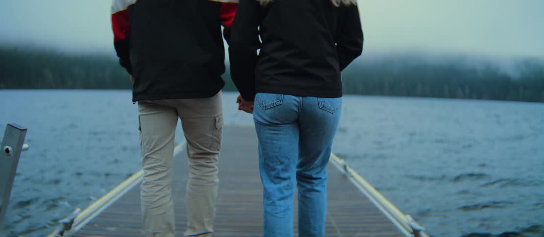 A young couple holds hands as they walk down a floating dock towards a lake on a cool, overcast day