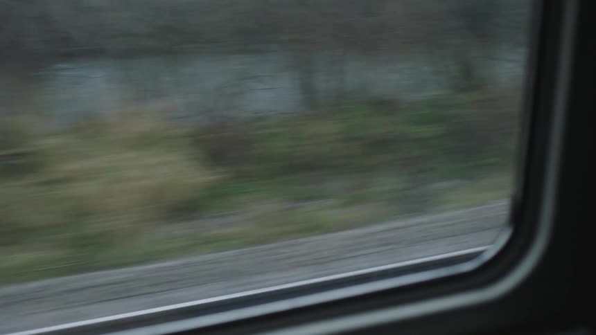 A view from a train window shows the blurry countryside and a river passing by on a gloomy, overcast day
