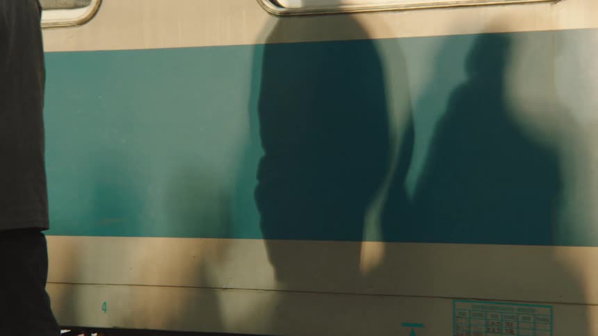 A slow-motion shot captures passengers waiting on a platform as their train begins to depart, casting long shadows in the sunlight