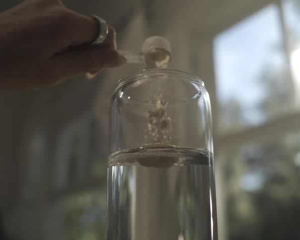 A close-up, slow-motion shot captures a hand pouring a scoop of supplement powder into a glass of clear water
