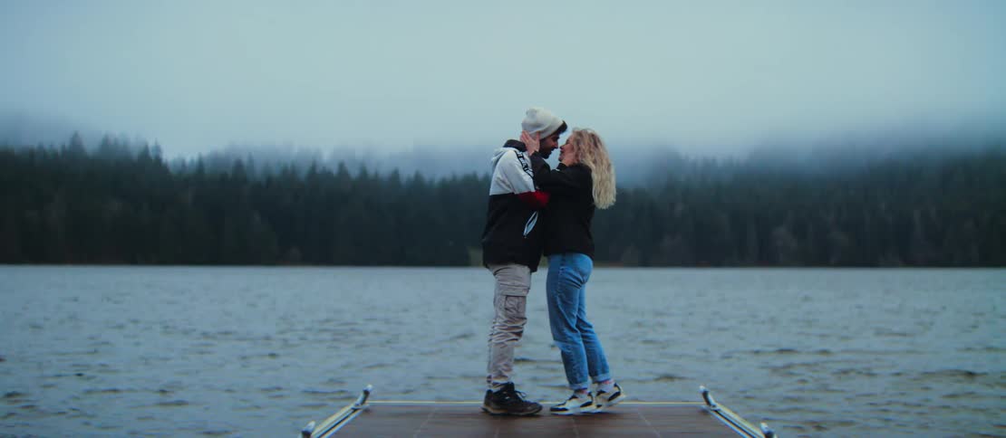 A young man surprises his girlfriend by getting down on one knee and proposing with an engagement ring on a dock overlooking a foggy lake