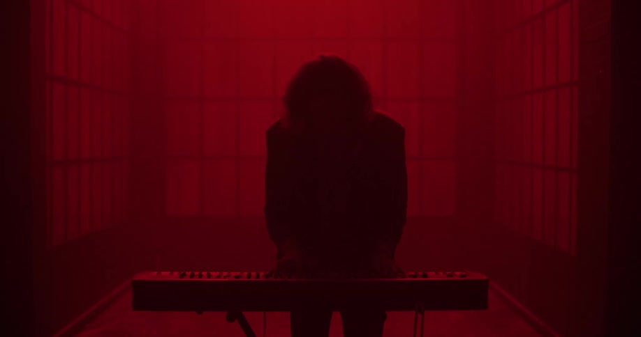 A silhouetted musician passionately plays a synthesizer keyboard in a dark, hazy room dramatically lit in red