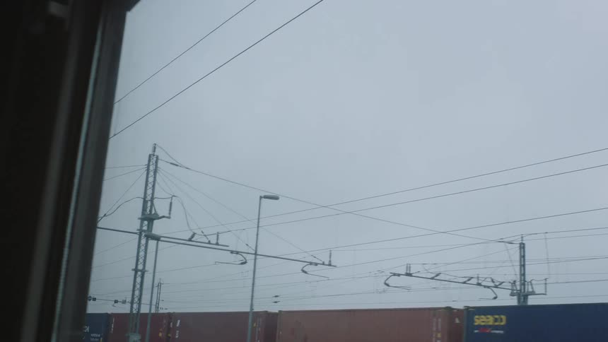 A slow-motion view from a train window passing by an industrial railyard with cargo containers on a grey, overcast day