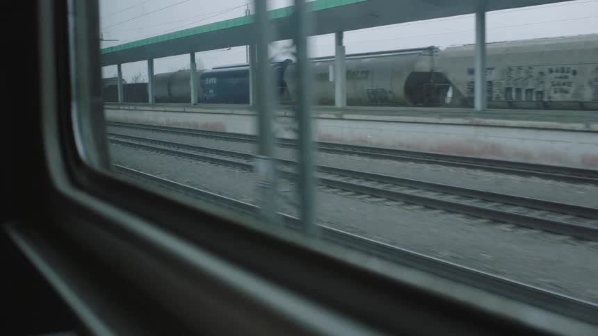 A view from a moving train window looking out at a freight train at a station on an overcast day