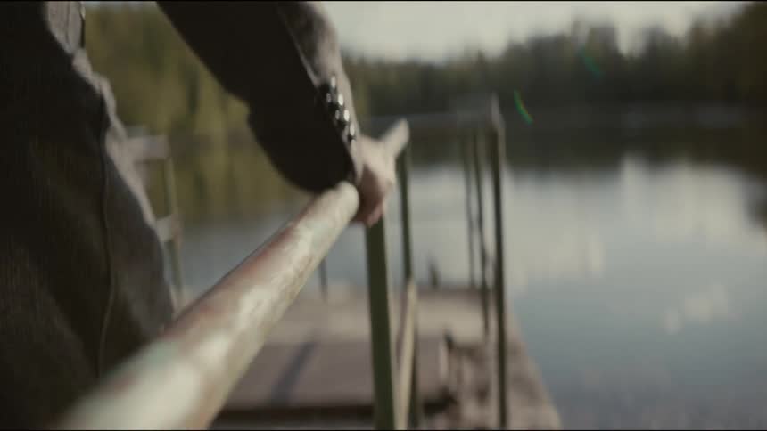 A man in a tweed jacket slowly walks along a pier, his hand gliding over the old metal railing in a pensive slow-motion shot