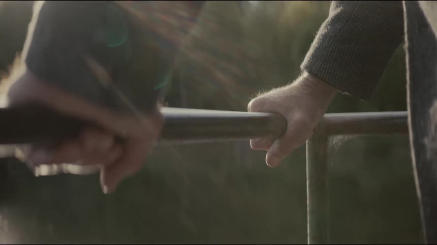 A close-up of two men's hands resting on a metal railing overlooking a calm river, bathed in the warm glow of the sun