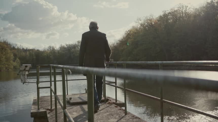 A senior man in a coat walks in slow motion to the end of a pier, looking out over a sun-dappled lake