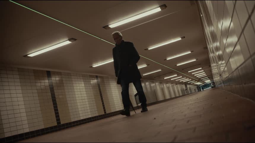 A well-dressed senior man walks slowly through a long, sterile pedestrian tunnel, leaning on his umbrella