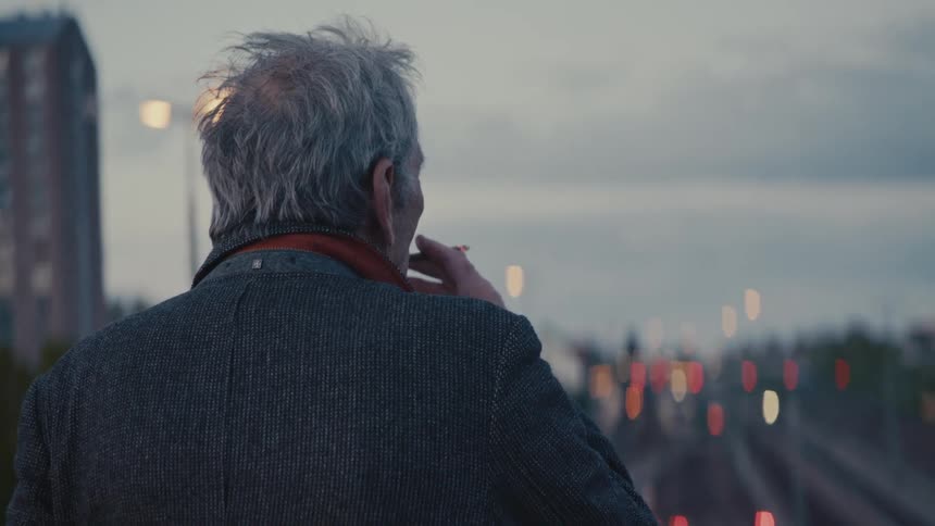 An older man smokes a cigarette in slow motion as he stands on a bridge looking out over the city lights at dusk