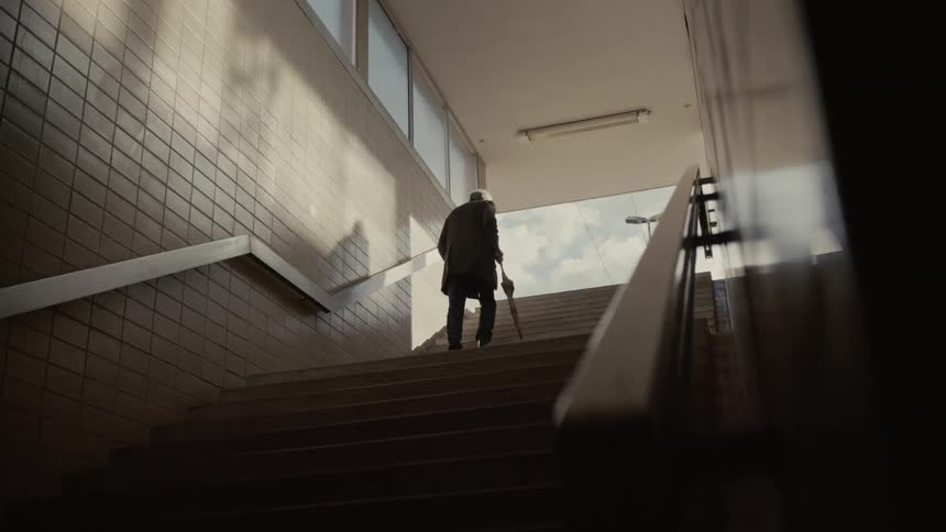 A silhouetted elderly man with an umbrella slowly climbs a long flight of stairs, emerging from a dark underpass into the light