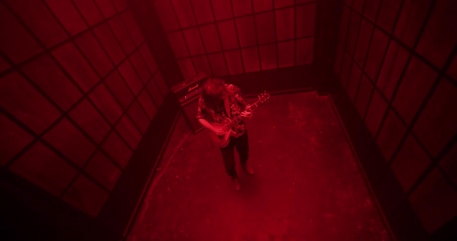 A long-haired guitarist shreds on an electric guitar in a moody, red-lit room with gridded walls, shot from a high angle