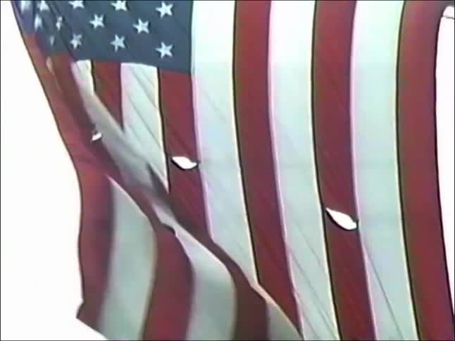 A tattered American flag waves in the wind, with the camera zooming out from a close-up to reveal the bustling street scene of Fifth Avenue in New York City