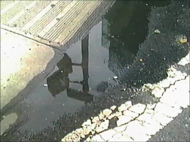 A person's foot in a sneaker steps into a dirty puddle on a city street, splashing muddy water