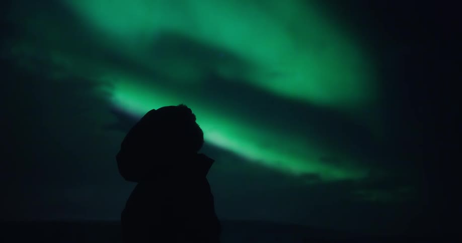 Cinemagraph of a silhouetted person watching the vibrant green Northern Lights, the Aurora Borealis, dance in the dark night sky