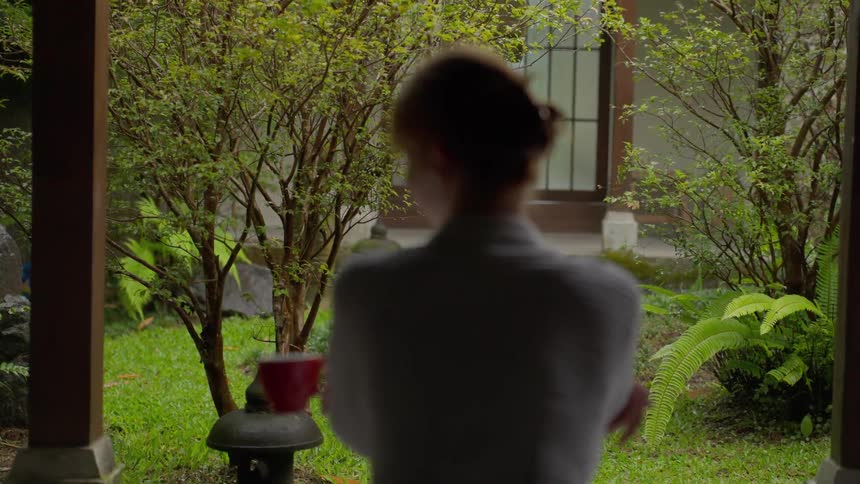 A woman in a cozy bathrobe starts her morning with a warm drink, looking out at a tranquil Japanese-style garden