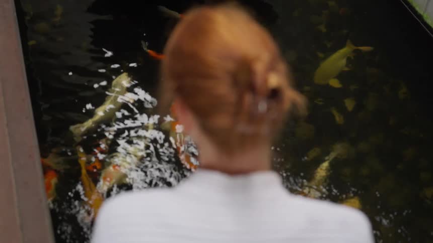 From over her shoulder, a woman with red hair watches as vibrant koi fish gracefully swim through a dark, tranquil pond