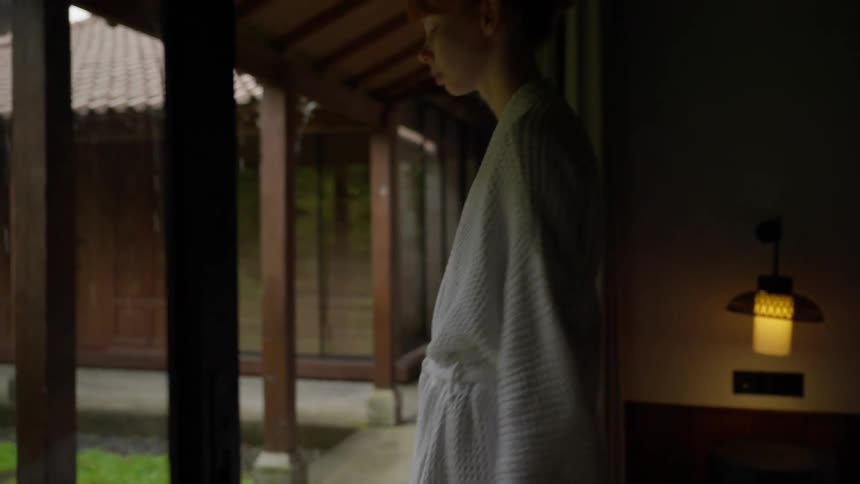 A woman in a bathrobe opens a sliding glass door and steps onto a patio to watch the rain in a peaceful garden