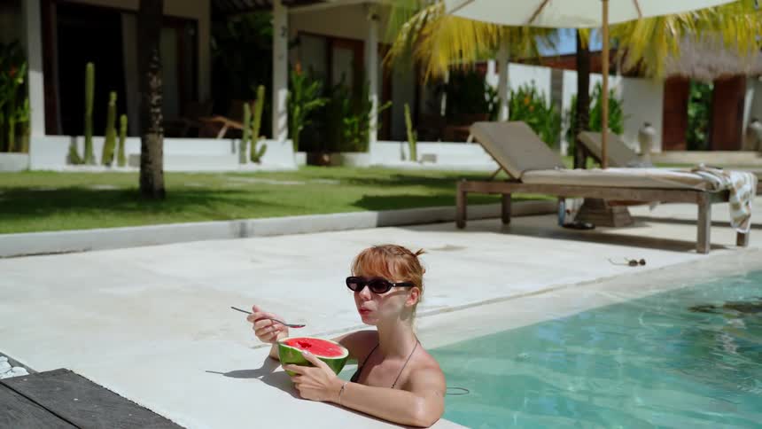 A woman in sunglasses enjoys a slice of watermelon with a spoon while relaxing in a swimming pool at a tropical resort in slow motion