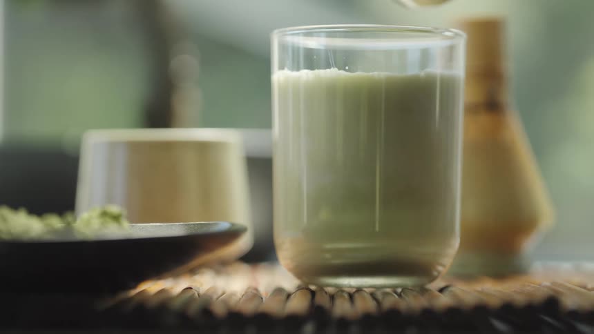 A close-up slow-motion shot captures the moment green matcha tea is poured into a glass of fresh milk to prepare a healthy and delicious matcha latte