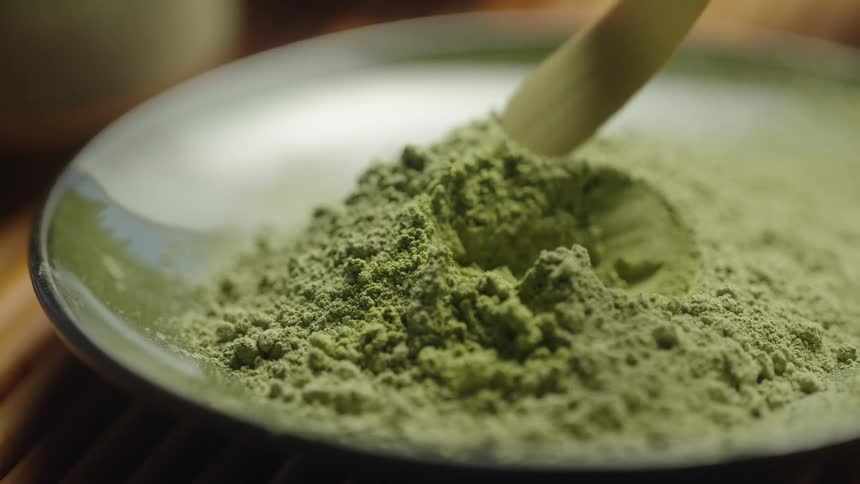 A wooden spoon gently scoops a serving of vibrant matcha green tea powder from a ceramic bowl in slow motion